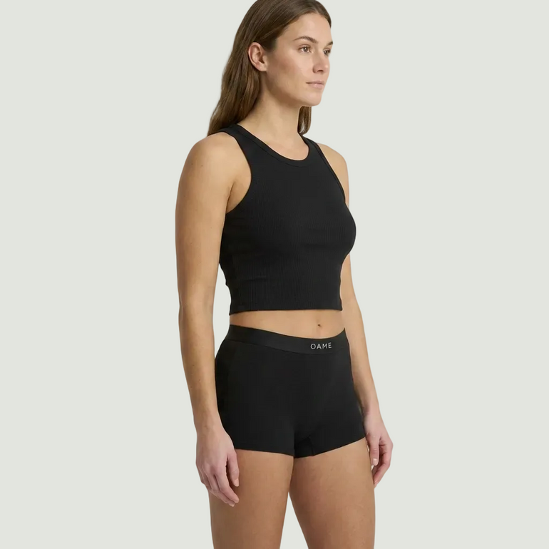 A woman modeling a black sleeveless crop top and matching black briefs against a plain light gray studio background. The brand name 'OAH' is visible on the waistband of the briefs.