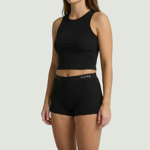 A woman modeling a black ribbed athletic crop top and matching black high-waisted shorts with 'DAME' printed in white on the waistband, set against a plain light gray studio background.