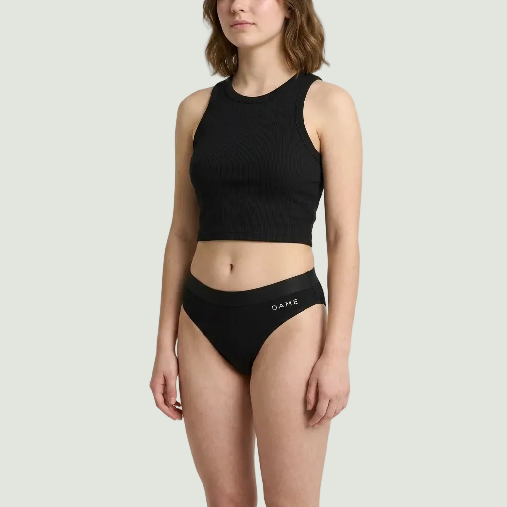 A model wearing a black ribbed crop top and matching black high-waisted briefs with the brand name 'DAME' printed in white on the front waistband, set against a plain light gray studio background.
