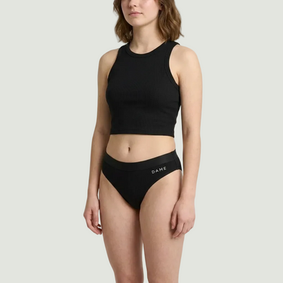 A model wearing a black ribbed crop top and matching black high-waisted briefs with the brand name 'DAME' printed in white on the front waistband, set against a plain light gray studio background.