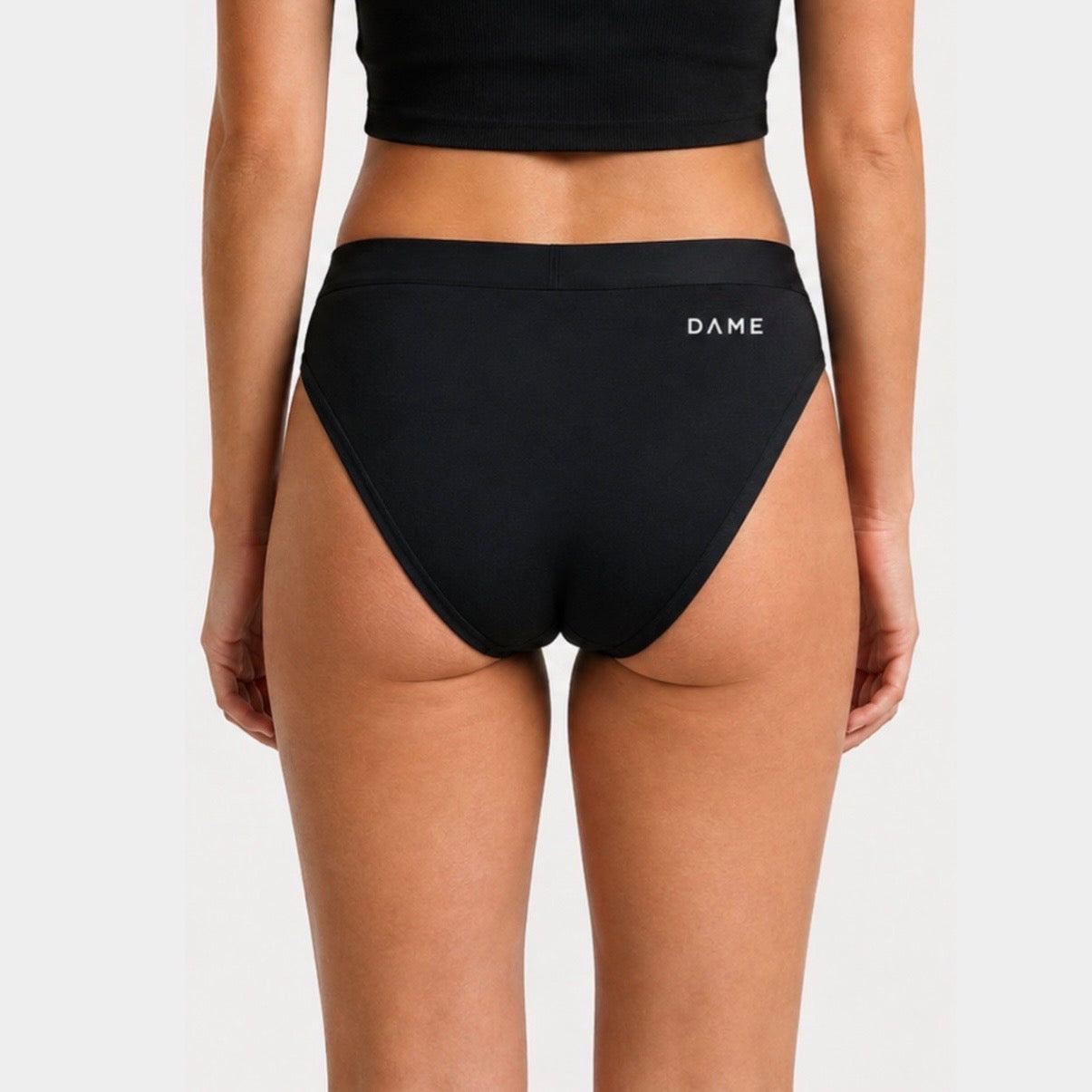 Rear view of a model wearing black high-cut bikini bottoms with the brand name 'DAME' printed in white on the back waistband, against a light gray studio background.