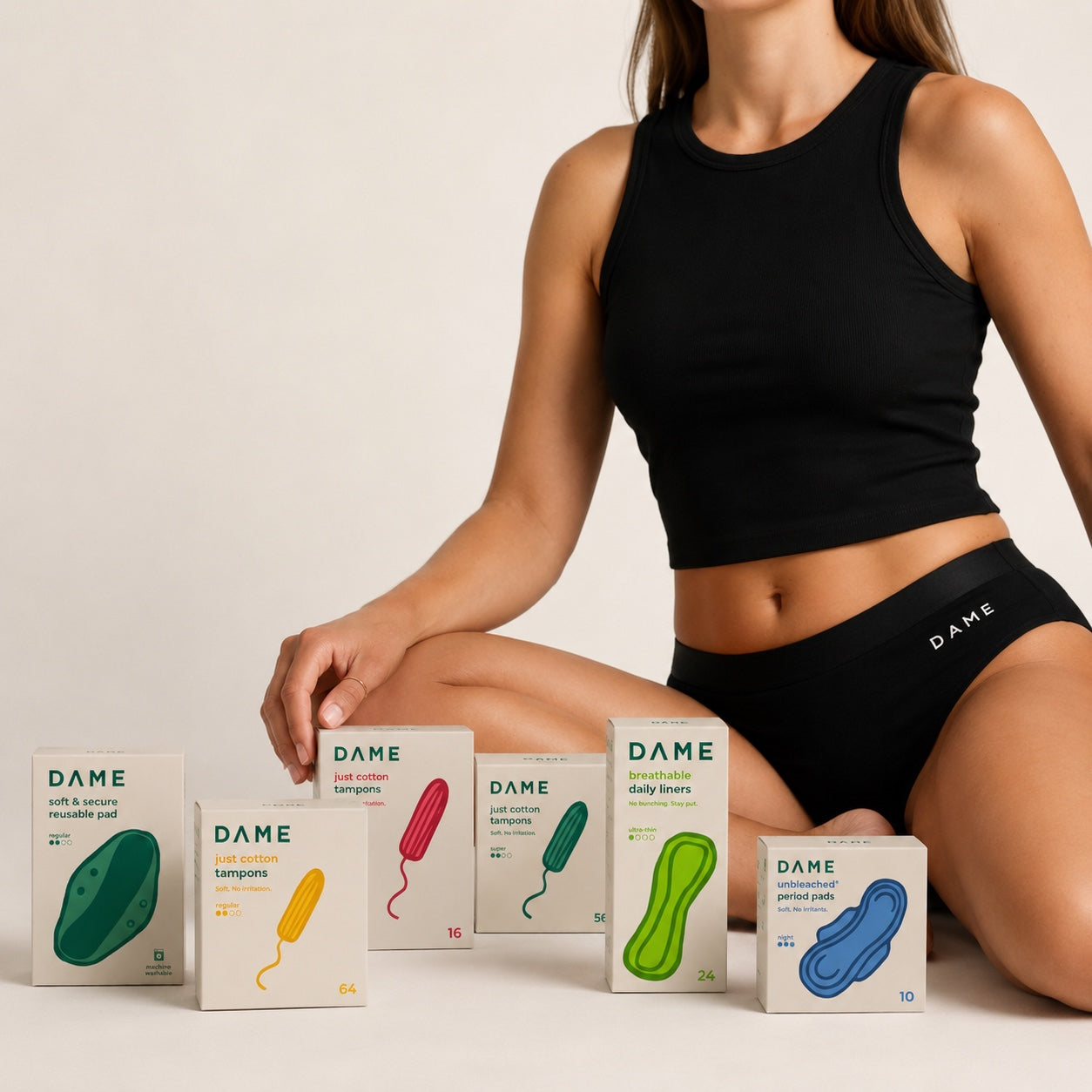 A woman in a black athletic crop top and shorts sits on a light beige studio floor, surrounded by several DAME branded boxes of menstrual pads and tampons. The boxes show different product colors including green, yellow, pink, teal, and blue.
