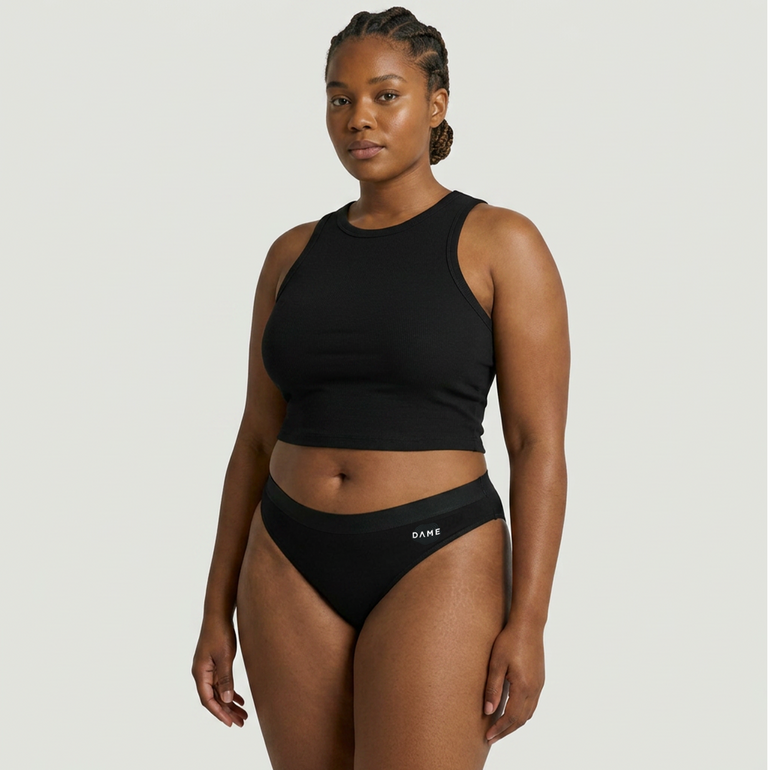 A woman modeling a black two-piece activewear set consisting of a sleeveless crop top and high-waisted briefs with a small white 'DANE' logo on the waistband, set against a solid light gray background.