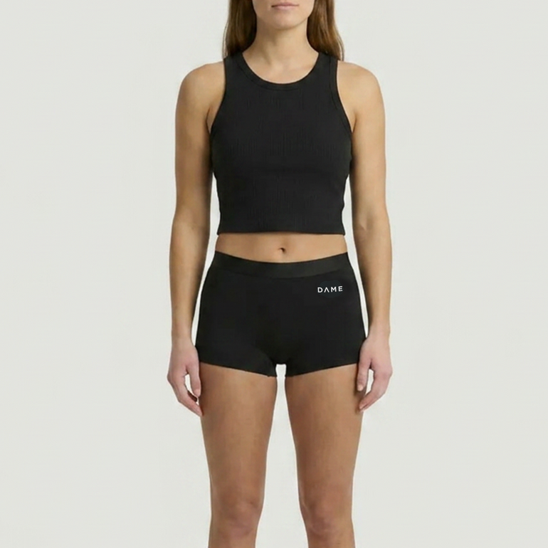 A woman modeling a black ribbed athletic crop top and matching black high-waisted briefs with a small white DAME logo on the waistband, set against a solid light gray background.