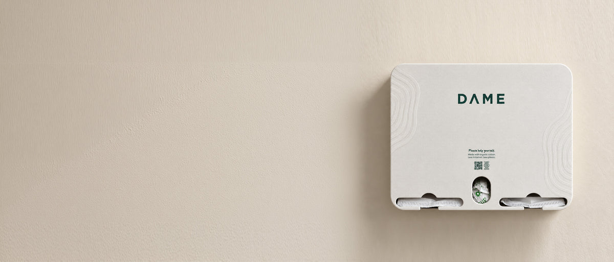 A white rectangular wall-mounted device with the brand name DAME printed in green on the top, shown against a plain beige wall.
